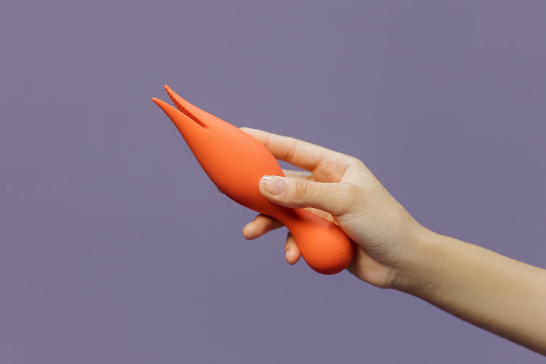 Close-up of a hand holding a modern clitoral vibrator against a minimalistic purple backdrop, symbolizing partnered pleasure and intimacy.