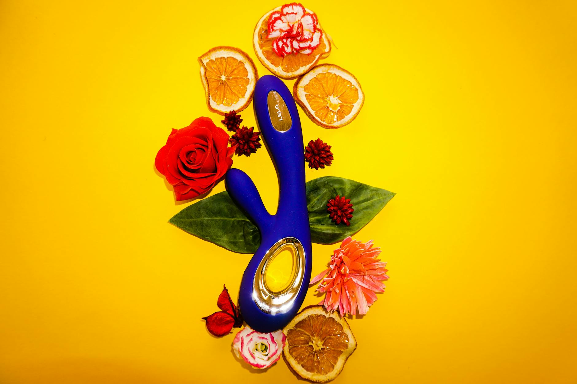 A blue clitoral vibrator surrounded by flowers and citrus slices on a bright yellow background